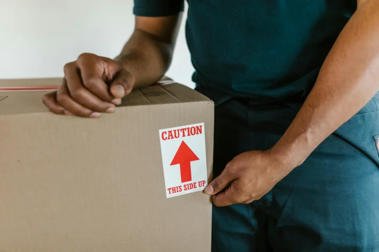 Primer plano de un operario de mudanzas colocando una etiqueta de "Caution This Side Up" en una caja de cartón para asegurar su transporte correcto.